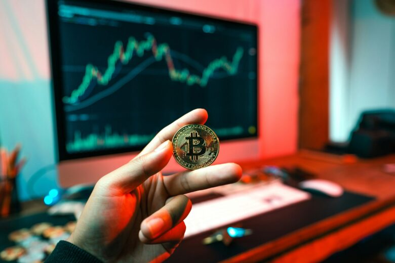 Tangan memegang koin Bitcoin di depan layar komputer dengan grafik naik.