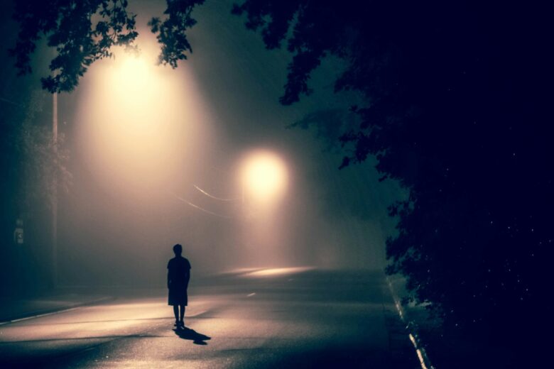 Sosok berdiri di tengah jalan yang berkabut di malam hari dengan lampu jalan menyala.