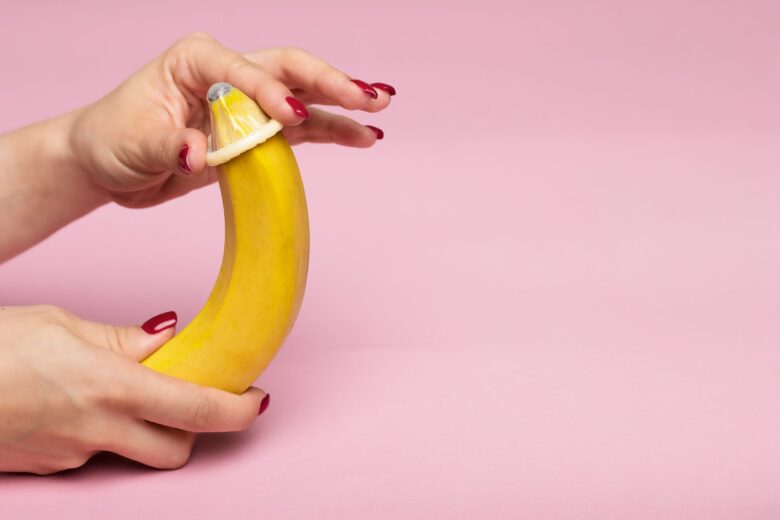 Tangan wanita mengaplikasikan kondom pada pisang kuning di latar belakang berwarna pink.