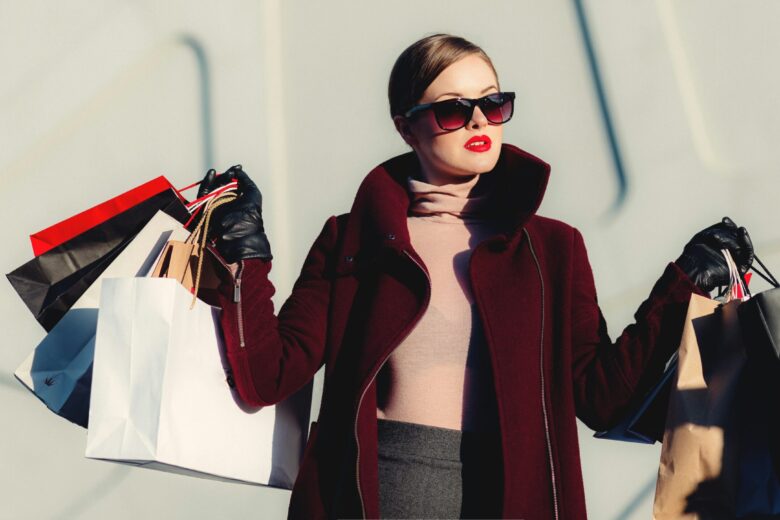 Wanita stylish mengenakan kacamata hitam, berdiri dengan beberapa tas belanja di tangan.