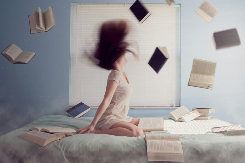Seorang wanita bergaun putih duduk di tempat tidur dengan buku-buku beterbangan di sekelilingnya. Rambutnya disanggul dan dia tersenyum.