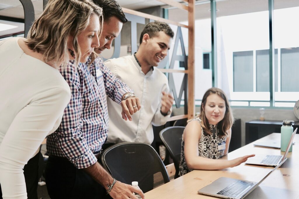 Sebuah tim yang terdiri dari empat orang berdiskusi di kantor. Dua orang duduk dan dua orang berdiri di belakang mereka. Mereka tampak terlibat dalam percakapan yang serius.