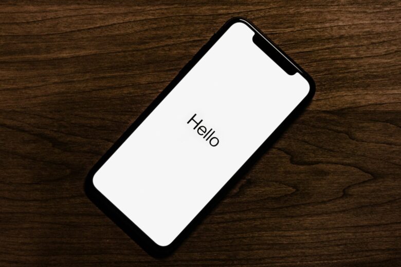 Sebuah Iphone berwarna hitam yang menampilkan kata "Halo" pada layar putih diposisikan secara diagonal pada permukaan kayu berwarna gelap.