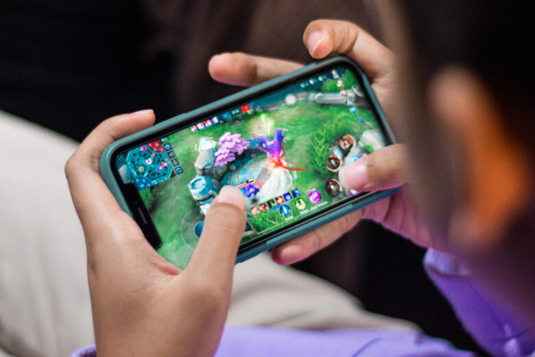 Seorang anak bermain game mobile dengan serius di smartphone, dengan layar yang menampilkan pemandangan permainan yang menarik.