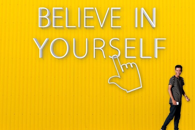 Seorang pria tersenyum di depan dinding kuning dengan tulisan "Believe in Yourself."