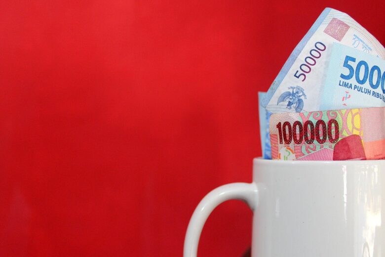 Cangkir putih berisi uang kertas Indonesia, termasuk 100.000 dan 50.000 rupiah, dengan latar belakang merah.