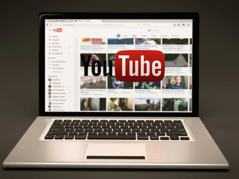 Laptop dengan situs web YouTube terbuka di layar. Logo YouTube berwarna merah tampak menonjol di bagian tengah layar.