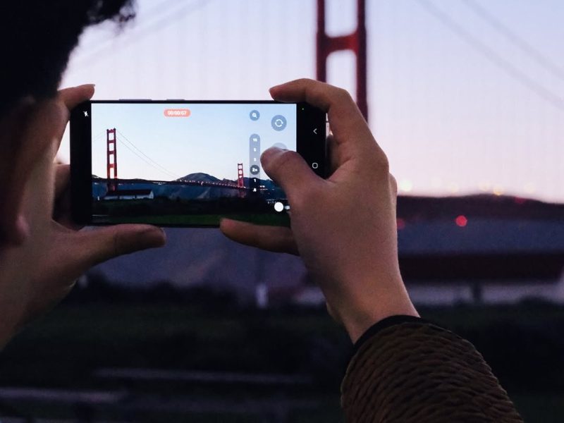 Seseorang mengambil foto Jembatan Golden Gate di San Francisco, California, dengan Samsung Galaxy S25 Ultra. Orang tersebut memegang ponsel dengan tangan kanannya, dan mereka sedang melihat ke layar. Jembatan Golden Gate terlihat di latar belakang, dan langit berwarna biru muda.