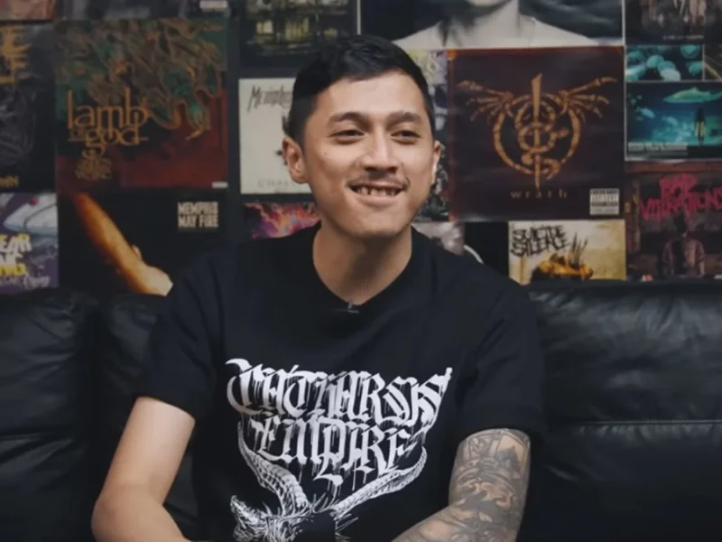 Bimo PD tersenyum mengenakan kaos hitam dengan desain metal, duduk di sofa dengan koleksi album musik metal di dinding.