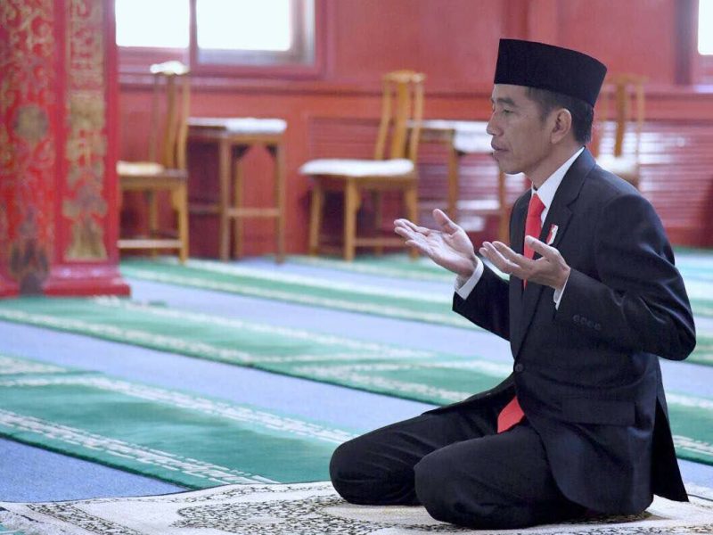 Joko Widodo sedang berdoa di dalam ruangan yang tenang, mengenakan jas formal dan peci.