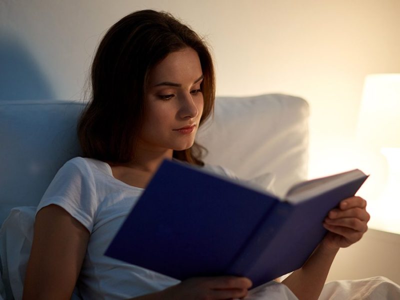 Gadis sedang membaca buku di tempat tidur dengan lampu hangat di sampingnya.