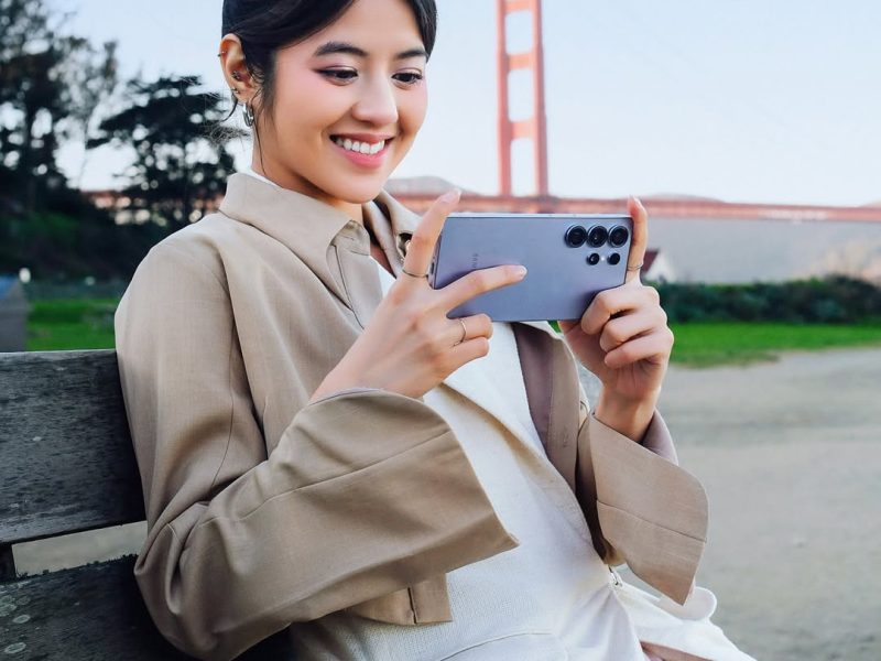 Seorang wanita mengenakan blazer cokelat dan kemeja putih duduk di bangku, tersenyum sambil memegang Samsung Galaxy S25 Ultra di tangannya. Dia sedang melihat ponsel, dan Jembatan Golden Gate dapat terlihat di latar belakang.