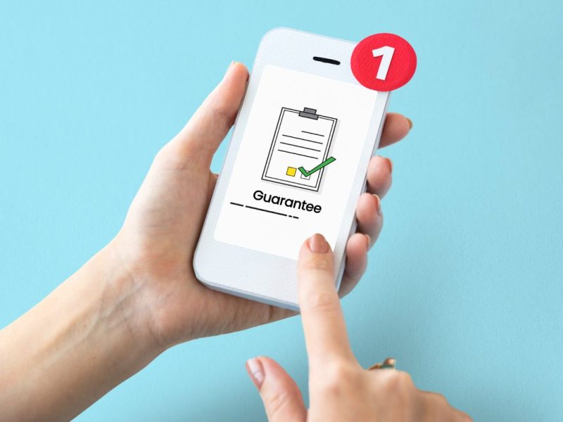 Tangan seseorang memegang telepon pintar dengan lencana notifikasi, yang menunjukkan tanda centang pada ikon dokumen dan kata "Jaminan".
