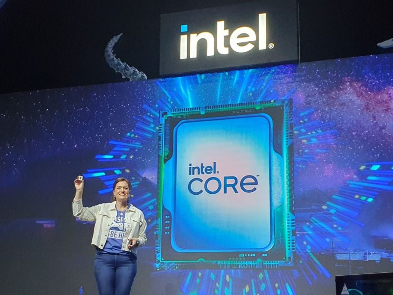 Seorang wanita dengan blazer krem tersenyum sambil mempresentasikan prosesor Intel Core. Tulisan "13th Gen D" terlihat di latar belakang.