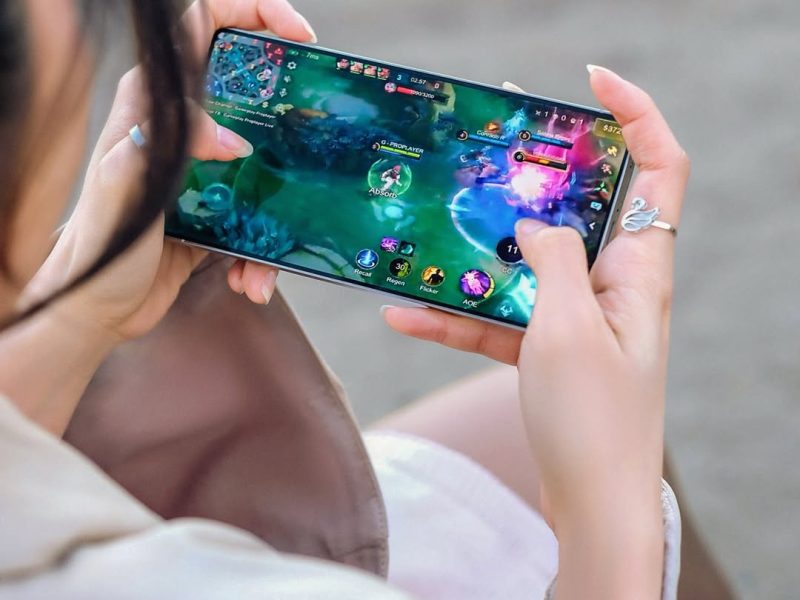 Seorang wanita muda duduk di luar dan bermain game mobile di ponselnya. Tangan kirinya memegang ponsel sementara tangan kanannya mengetuk layar. Antarmuka game menampilkan mobile legends. Dia mengenakan gaun putih dan jaket cokelat.