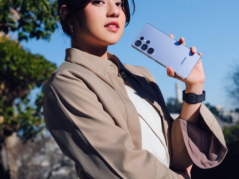 Seorang wanita muda dengan rambut hitam memegang Samsung Galaxy S22 Ultra berwarna lavender. Ia mengenakan blazer berwarna beige muda di atas kemeja putih. Foto diambil di luar ruangan, dengan langit biru dan tanaman hijau di latar belakang.