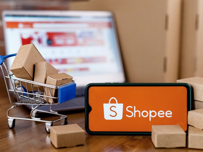 Gambar smartphone menampilkan aplikasi Shopee dengan keranjang belanja kecil dan kotak paket di latar belakang.