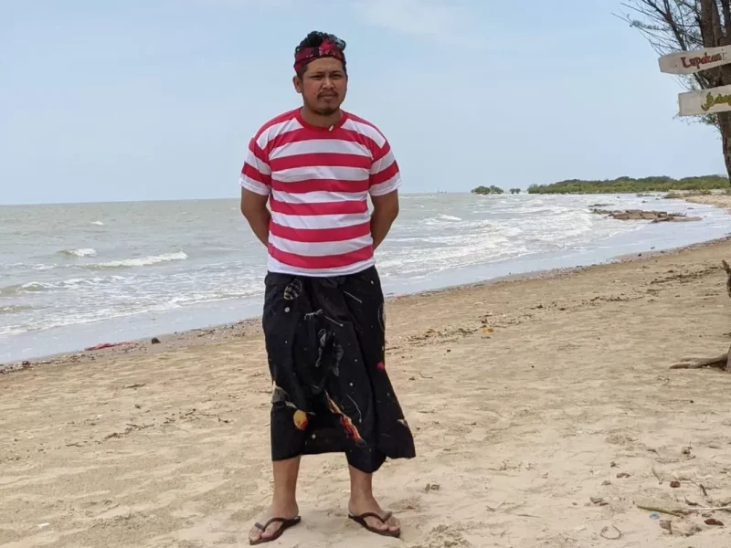 Pria berdiri di tepi pantai mengenakan kaos bergaris merah-putih dan kain sasirangan hitam.