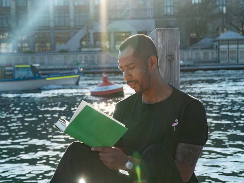 Pria sedang membaca buku di tepi sungai dengan latar belakang perahu dan sinar matahari.