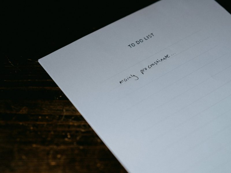 Kertas daftar tugas dengan tulisan 'mainly procrastinate' di atas meja kayu gelap.