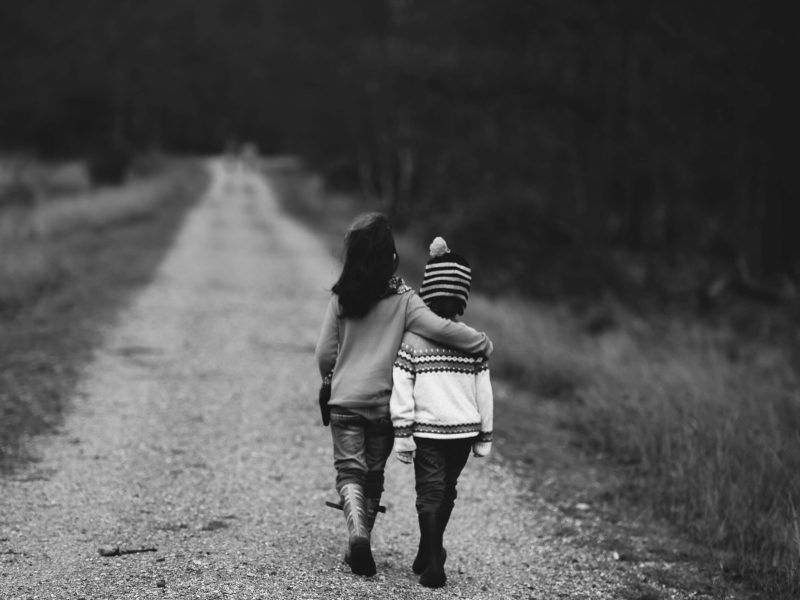 Dua anak berjalan berdampingan di jalan setapak yang dikelilingi pepohonan, tampak akrab dalam suasana hitam-putih.