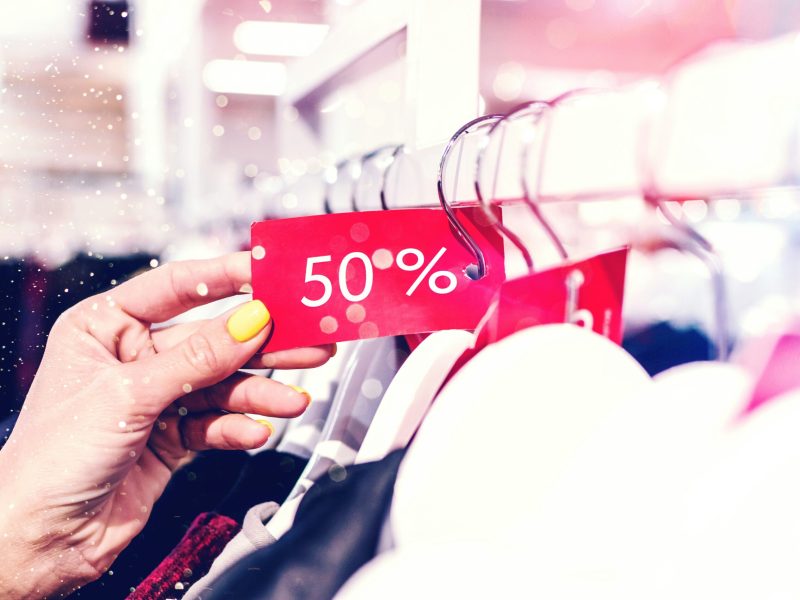 Obral Diskon 50%! Tangan memegang label merah bertuliskan diskon 50%, tergantung di rak pakaian di sebuah toko.