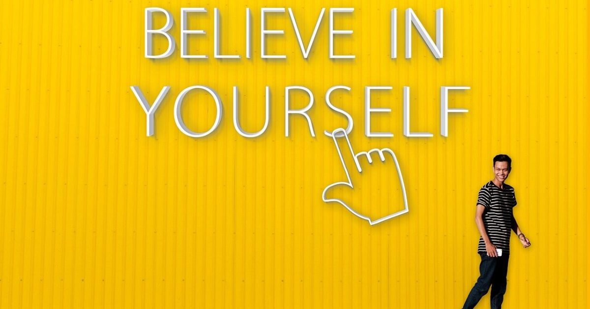 Seorang pria tersenyum di depan dinding kuning dengan tulisan "Believe in Yourself."