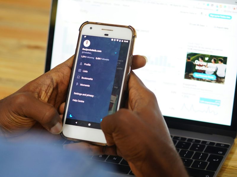 Pengguna Smartphone yang Mengakses Akun Media Sosial Seseorang menggunakan smartphone untuk mengakses akun media sosial, dengan laptop di latar belakang.