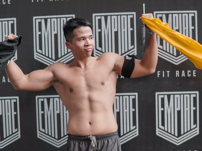 Seorang pemuda atletis menunjukkan ototnya di acara Empire Fit Race, mengenakan celana pendek dan mengangkat baju hitam serta kuning.