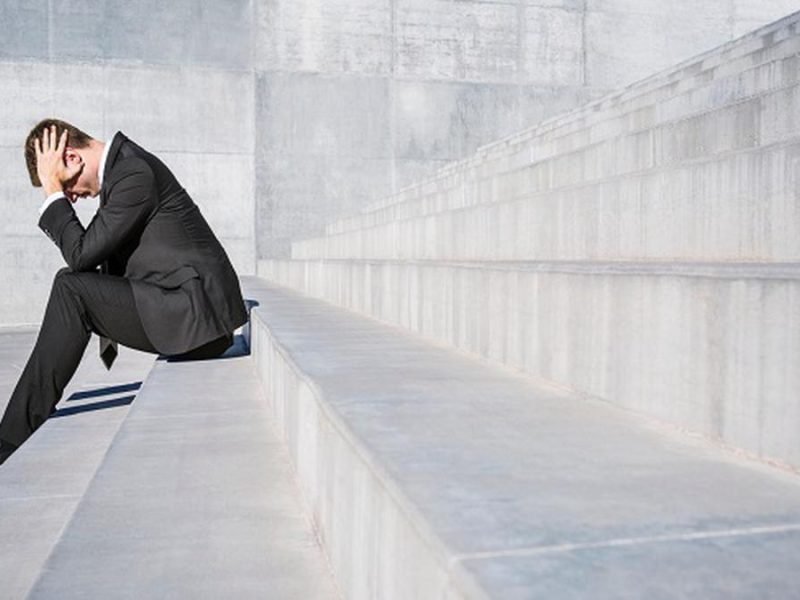 Pria mengenakan setelan formal duduk dengan tangan di kepala, mengekspresikan perasaan stress atau kesedihan di tangga beton.