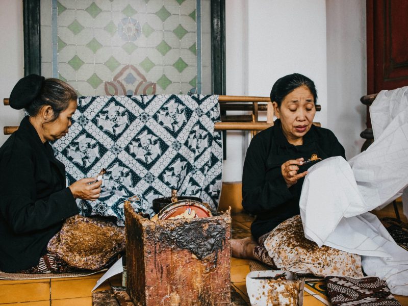 Dua pengrajin batik wanita sedang bekerja dengan alat tradisional di ruang kerajinan.