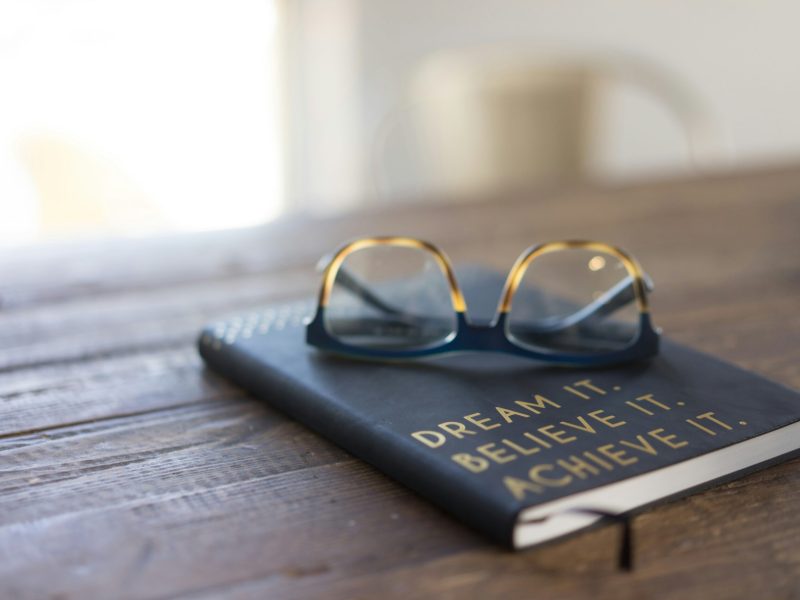 Buku Jurnal Dengan Kutipan Motivasi Sebuah jurnal hitam dengan tulisan emas bertuliskan "Dream It. Believe It. Achieve It." diletakkan di atas meja kayu. Kacamata diletakkan di atas jurnal tersebut.
