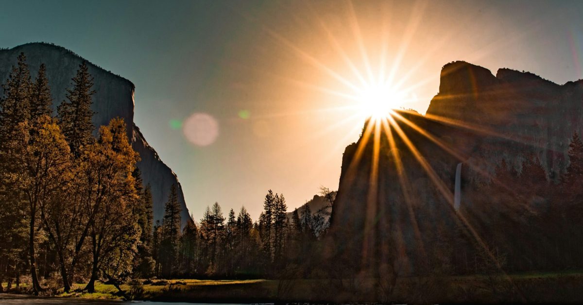 Matahari terbenam di atas pegunungan Yosemite National Park, dengan sinar matahari yang menyinari pohon-pohon dan sungai.