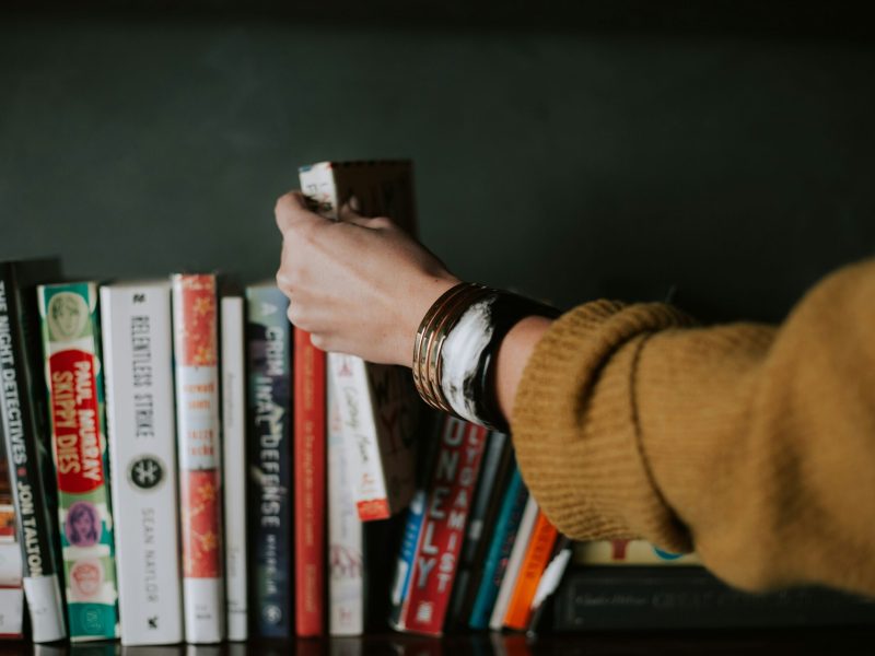 Seorang wanita dengan gelang emas mengambil buku dari rak penuh dengan berbagai judul.