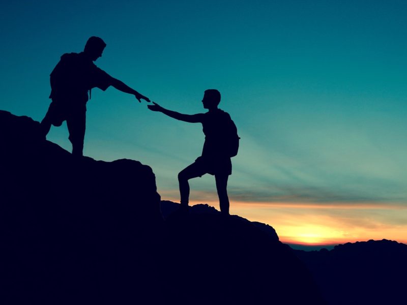 Silhouette dua pendaki gunung yang saling membantu dan berinteraksi di puncak gunung dengan latar belakang langit senja yang indah.