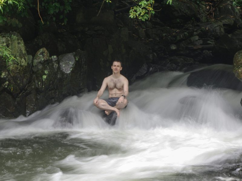 Meditasi di Aliran Air Pria sedang meditasi di atas batu yang dikelilingi aliran air.