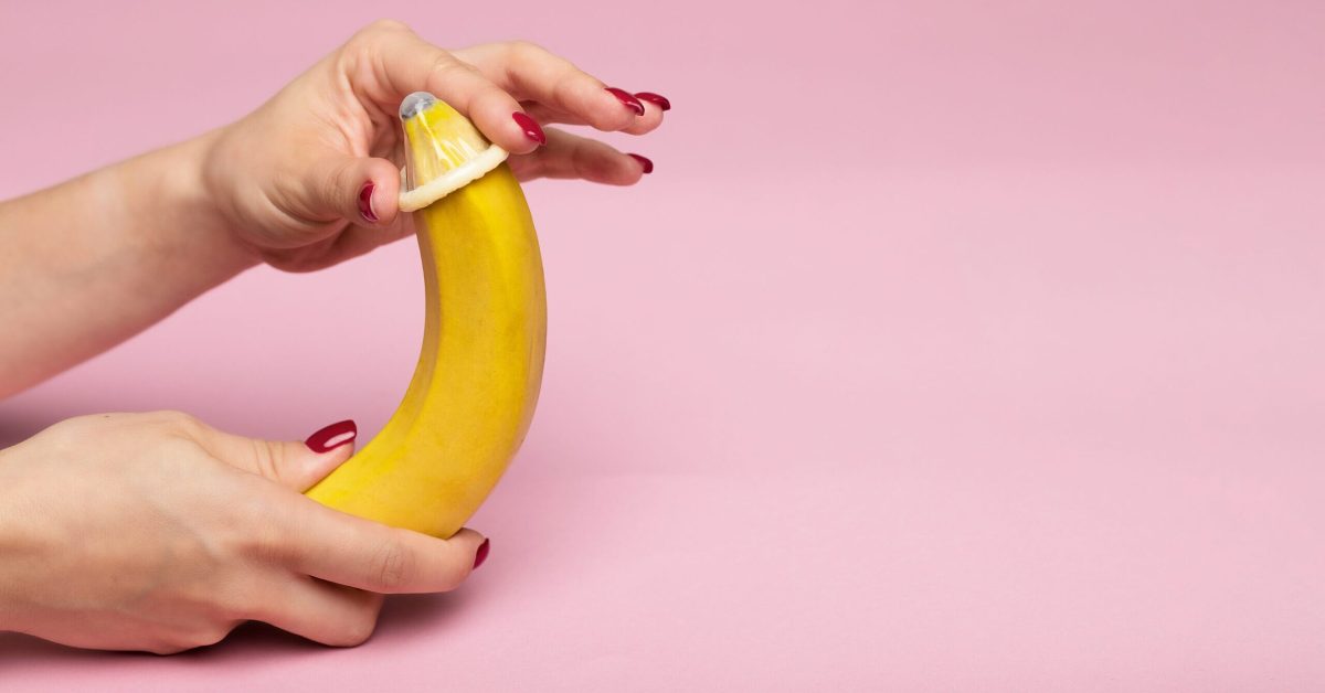 Pentingnya Edukasi Seksual: Menggunakan Kondom dengan Benar Tangan wanita mengaplikasikan kondom pada pisang kuning di latar belakang berwarna pink.