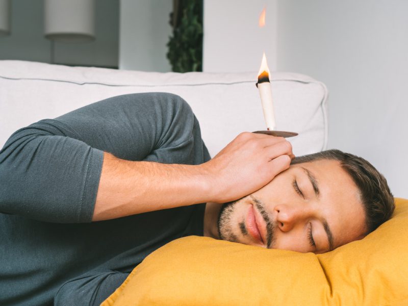 Ear Candling: Aman atau Tren yang Berbahaya? Seorang pria berbaring di atas bantal kuning dengan mata tertutup, lilin menyala dipegang dekat telinganya.