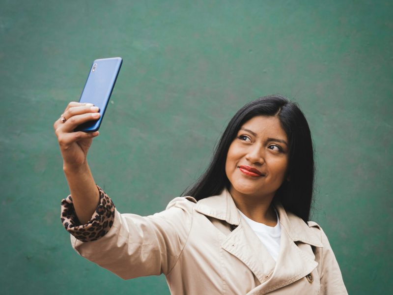 Seorang wanita muda berambut hitam panjang sedang berswafoto dengan telepon pintar biru. Ia mengenakan mantel panjang krem ​​dengan lapisan motif macan tutul. Wanita itu tersenyum dan melihat ke samping. Latar belakangnya adalah dinding hijau.