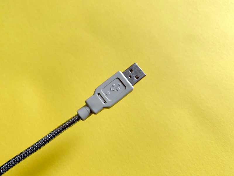 Foto close-up kabel USB putih dengan konektor fleksibel berwarna perak pada latar belakang kuning.