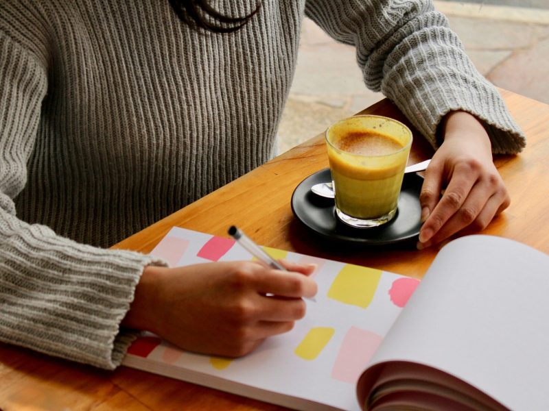Menulis di Notebook dengan Segelas Turmeric Latte Teks Alt Tangan seseorang menulis di buku catatan dengan pena sambil memegang secangkir latte kunyit.