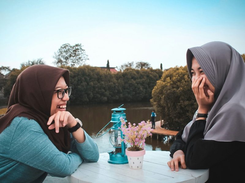 Dua wanita mengenakan hijab tersenyum saat berbincang di tepi danau, dengan lampu minyak tua dan tanaman hias di meja.