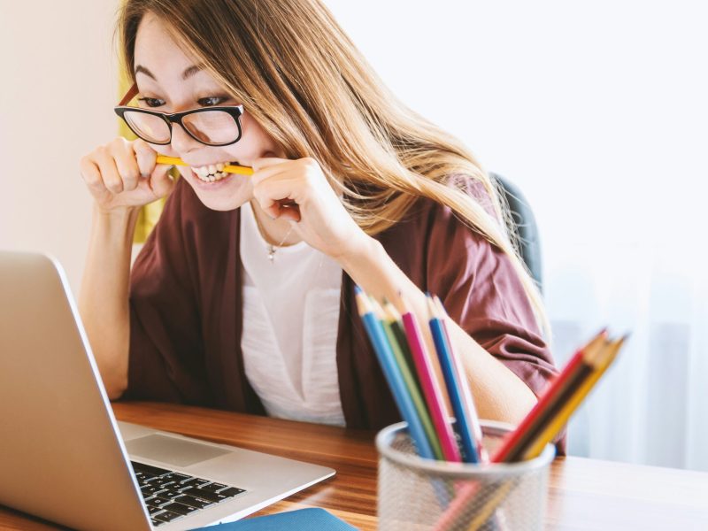 Wanita Tertekan Bekerja di Depan Laptop 158 / 5.000 Seorang wanita muda berkacamata menggigit pensil kuning sambil menatap laptop. Ia duduk di meja dengan secangkir pensil warna di depannya.
