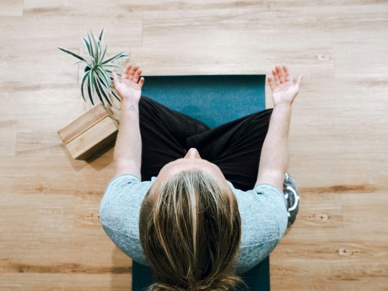 Seorang wanita melakukan yoga di lantai kayu Seorang wanita berambut cokelat bermeditasi dengan mata tertutup di atas matras yoga biru. Dia duduk bersila dengan kedua tangan terentang di sampingnya. Dia memakai kaus biru muda dan celana hitam. Dia bermeditasi di atas lantai kayu dengan tanaman hijau di belakangnya.