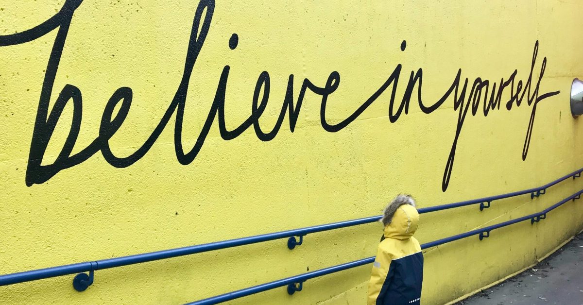 Anak Berdiri di Dekat Dinding “Percayalah pada Diri Sendiri” Seorang anak dengan jaket musim dingin kuning dan biru berdiri di dekat dinding kuning dengan kata-kata "percayalah pada dirimu sendiri" yang ditulis dengan cat hitam.
