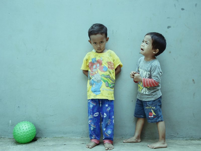 Dua anak laki-laki berdiri di depan dinding berwarna biru, satu tampak marah sementara yang lain menangis, dengan bola hijau di samping mereka.