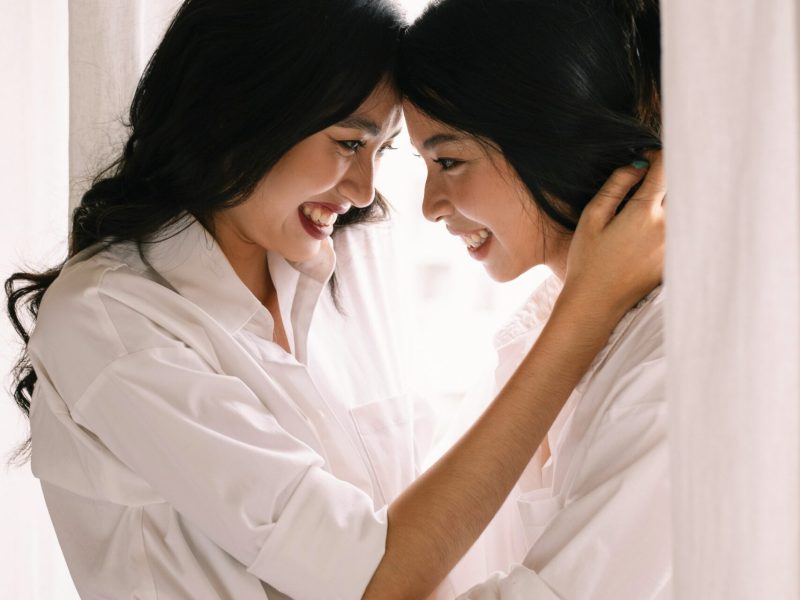 Wanita Tersenyum Berbaju Putih Dua wanita berbaju putih saling tersenyum, berdiri di ruangan terang dengan tirai putih.
