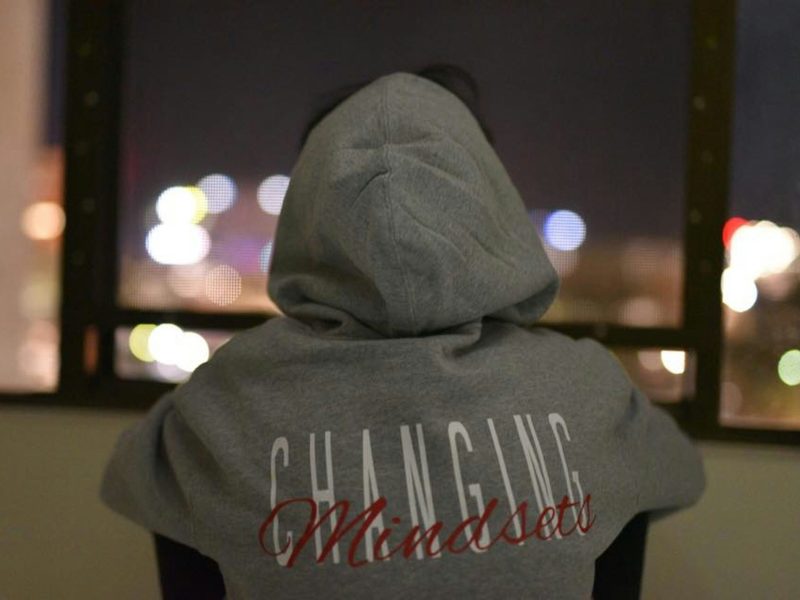 Seseorang Memakai Hoodie dengan Tulisan “Changing Mindsets” Gambar ini memperlihatkan seorang pria memakai hoodie abu-abu dengan tulisan "Changing Mindsets" di bagian belakang. Hoodie tersebut memiliki warna putih dan merah pada tulisannya. Dia duduk menghadap jendela yang memperlihatkan pemandangan kota di malam hari.