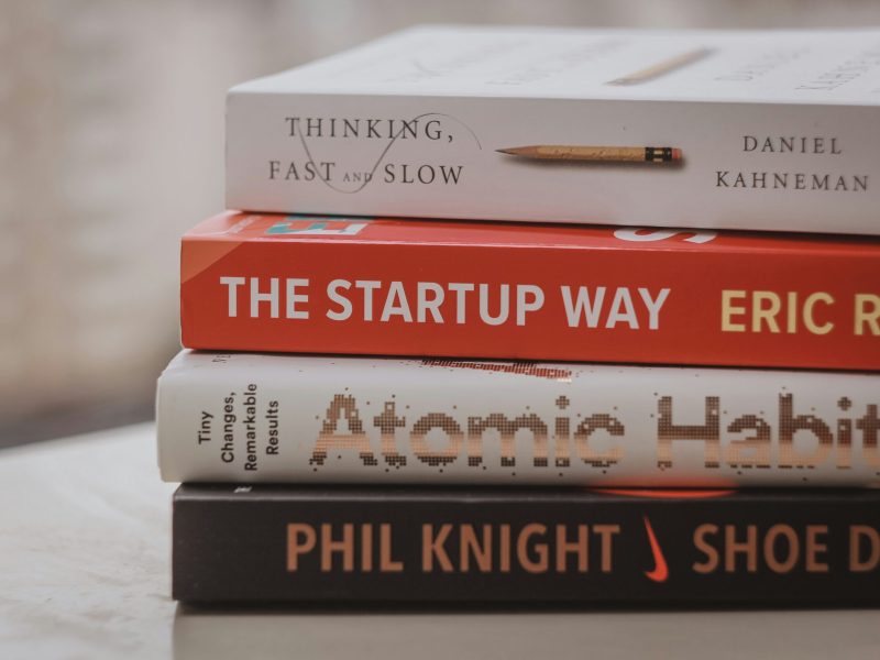 Tumpukan Buku tentang Pengembangan Diri dan Bisnis Tumpukan empat buku, masing-masing dengan judul berbeda. Dari atas ke bawah, buku-buku tersebut adalah "Thinking, Fast and Slow" karya Daniel Kahneman, "The Startup Way" karya Eric Ries, "Atomic Habits" karya James Clear, dan "Shoe Dog" karya Phil Knight.