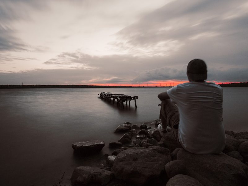 Seorang pria duduk di atas batu-batu di tepi danau, melihat ke arah dermaga kayu yang hancur di tengah danau. Langit berwarna merah muda dan abu-abu di cakrawala, dan permukaan air tampak tenang.